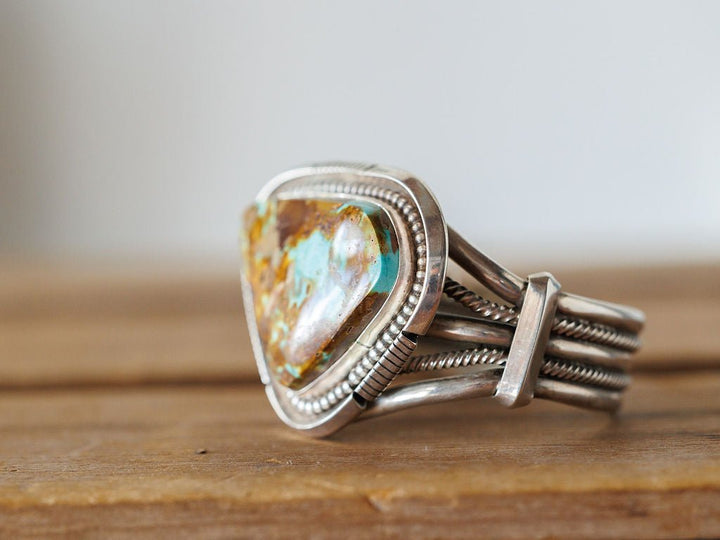 The Sedalia Cuff by Savannah Sevens western life{&}style features a large, unique turquoise and brown stone set in sterling silver, showcased on wood with a neutral backdrop—a striking piece of Native American-inspired jewelry.
