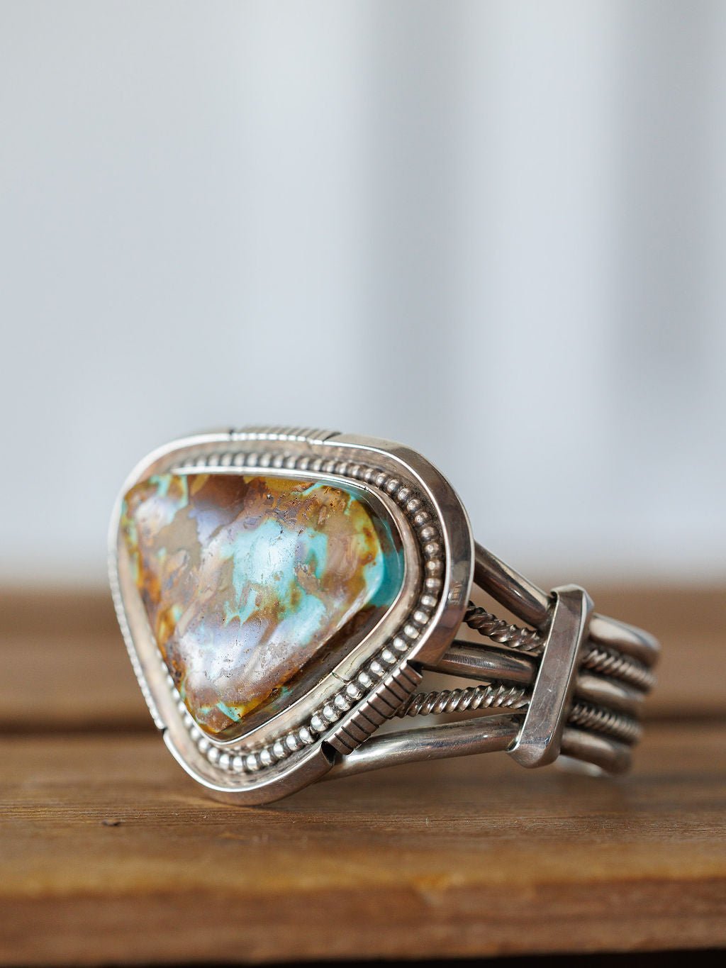 The Sedalia Cuff by Savannah Sevens western life{&}style is a sterling silver bracelet with a large, triangular turquoise stone featuring brown and blue marbling in an ornate, beaded frame, inspired by Native American jewelry and displayed on wood.