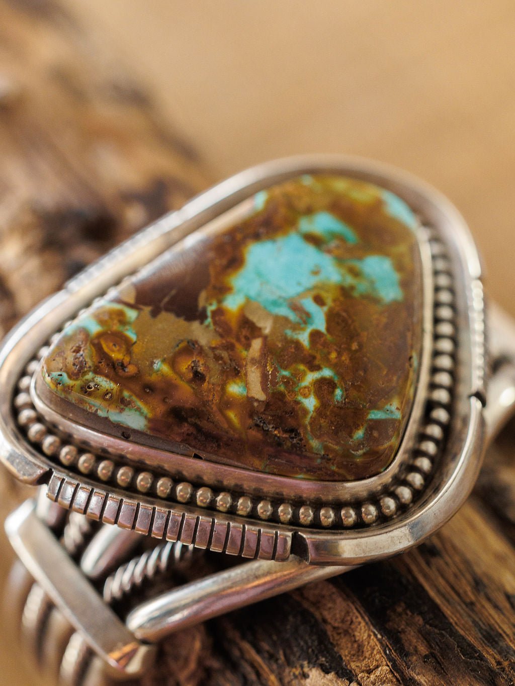 A close-up of the Sedalia Cuff by Savannah Sevens western life{&}style, showcasing a triangular turquoise and brown stone set in sterling silver with intricate details, inspired by Native American jewelry, on a rustic wooden surface.