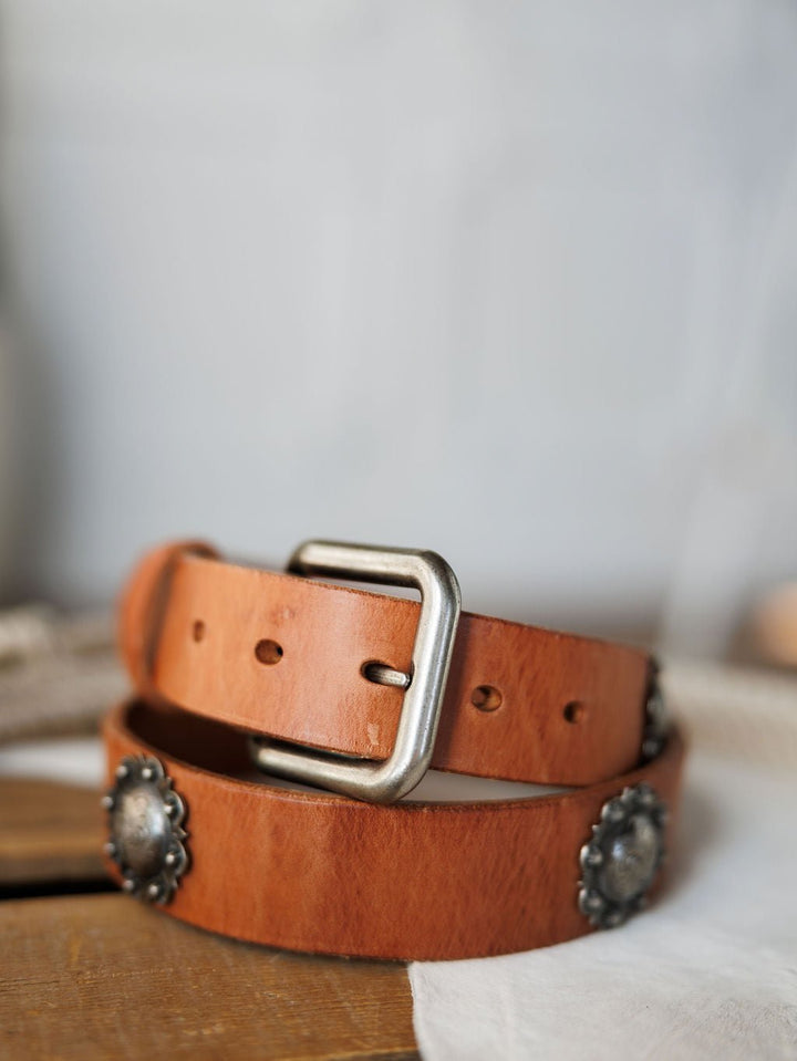 A close-up of the Sedona Belt by Savannah Sevens western life{&}style, featuring cognac leather, a silver buckle, and decorative southwest conchos, displayed on a light-colored fabric.