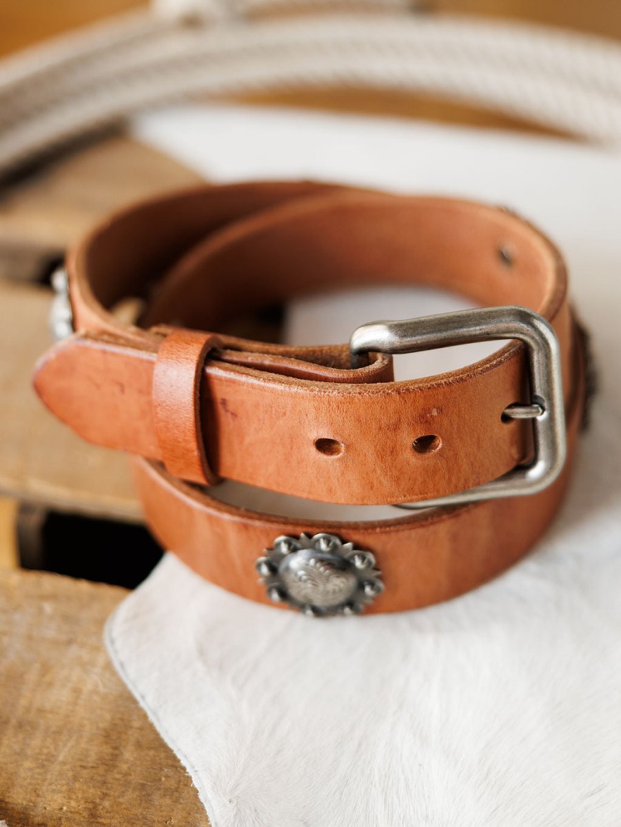The Sedona Belt by Savannah Sevens western life{&}style is a cognac leather western belt with southwest silver conchos and a silver buckle, coiled on a light surface with rope and wood pieces in the background.