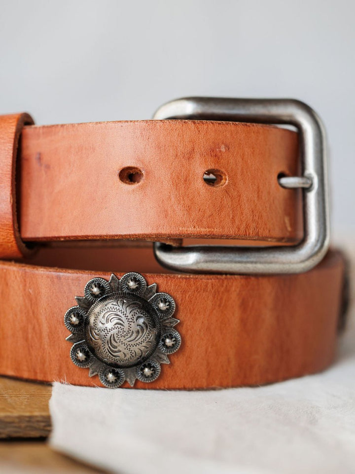 A close-up of the Sedona Belt by Savannah Sevens western life{&}style, featuring cognac leather, a silver rectangular buckle, and an engraved southwest concho with floral details, shown resting on white fabric.