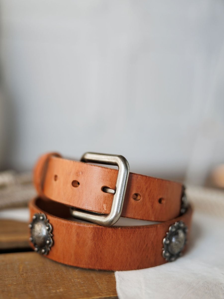 A close-up of the Sedona Belt by Savannah Sevens western life{&}style, featuring cognac leather, a silver buckle, and decorative southwest conchos, displayed on a light-colored fabric.