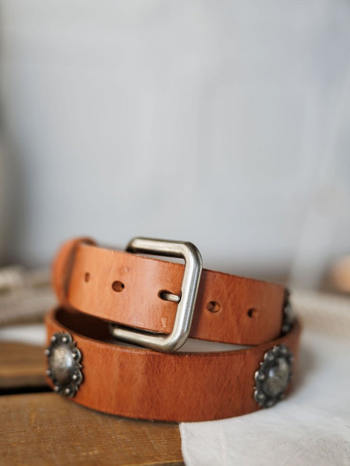 A close-up of the Sedona Belt by Savannah Sevens western life{&}style, featuring cognac leather, a silver buckle, and decorative southwest conchos, displayed on a light-colored fabric.