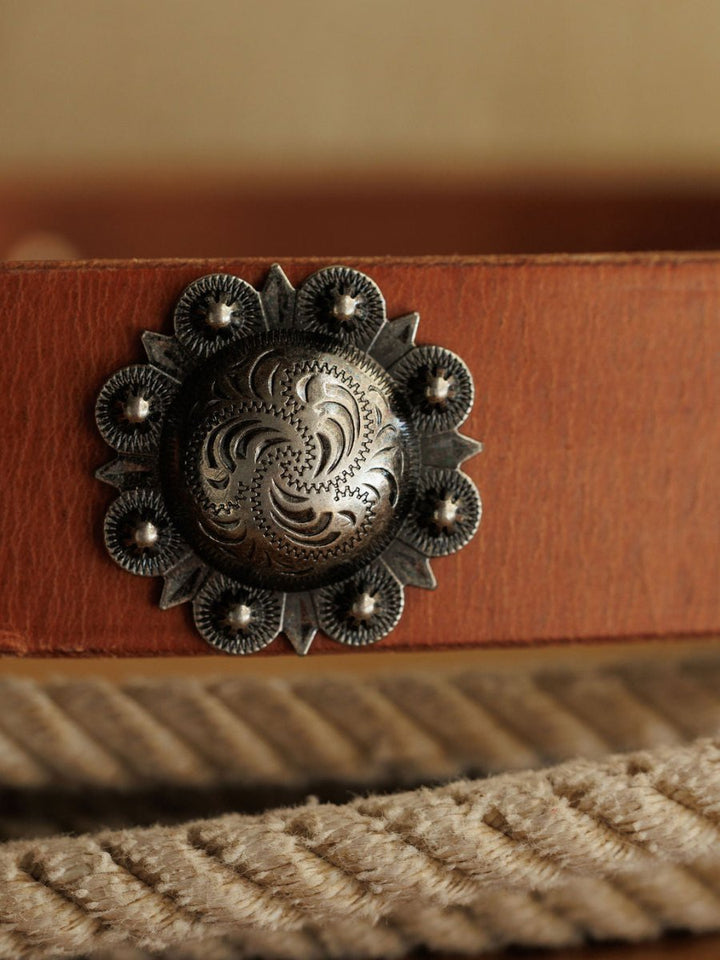 A close-up of a Savannah Sevens western life{&}style Sedona Belt in cognac leather with an ornate engraved southwest silver concho. A thick, coiled rope appears softly blurred in the background.