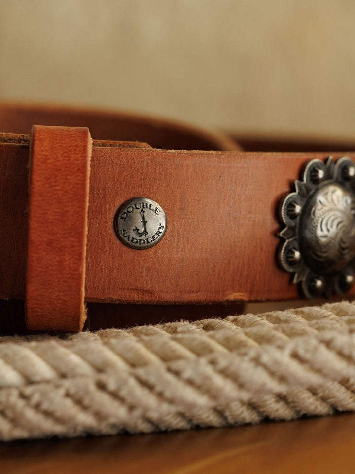 A close-up of the Sedona Belt by Savannah Sevens western life{&}style, crafted in cognac leather with a southwest silver concho and round metal tag, resting on coiled rope against a blurred background.