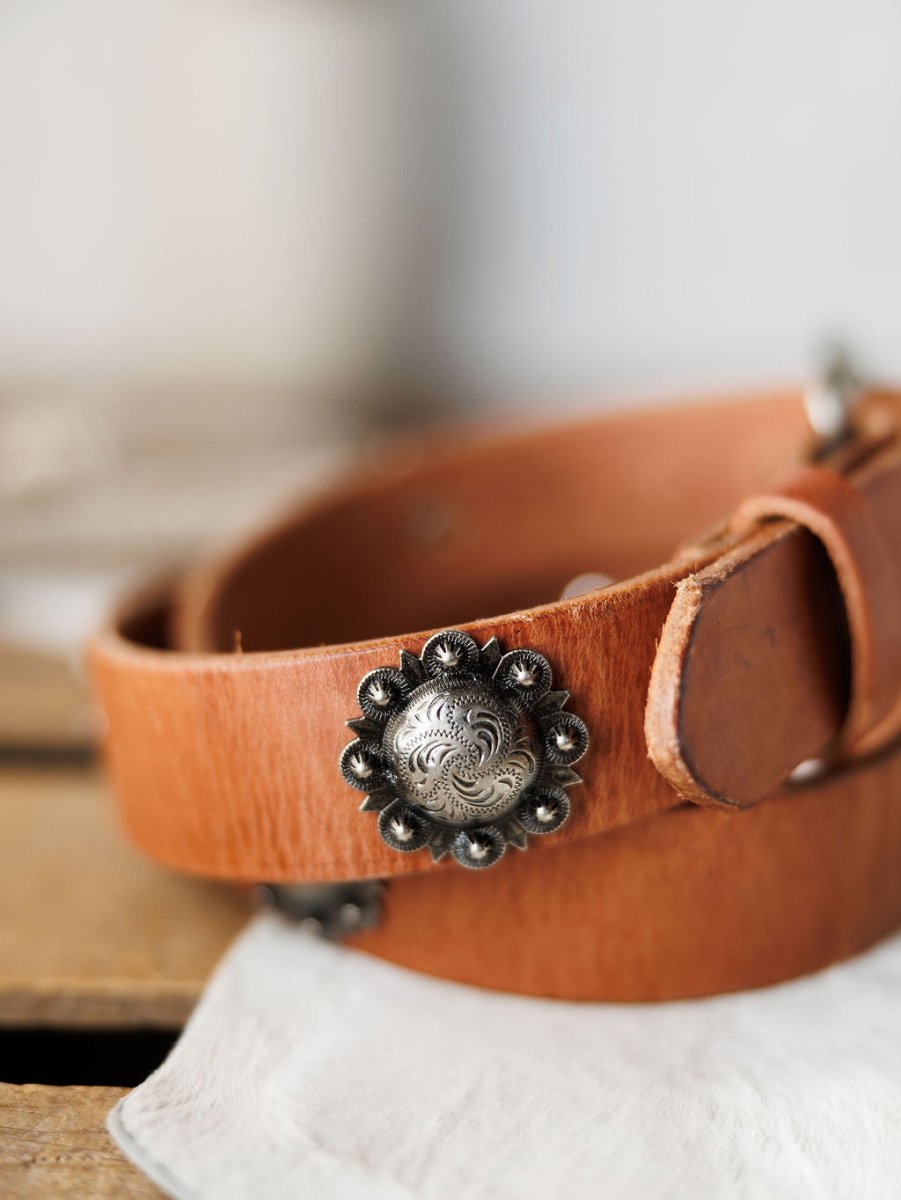 A close-up of the Sedona Belt by Savannah Sevens western life{&}style, showing a cognac leather western belt with an ornate southwest silver concho and floral designs, resting on a soft white cloth with a blurred background.