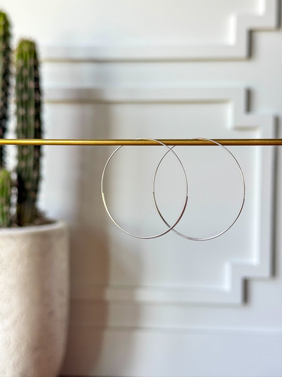 Two thin, 14K white gold plated Seminole Earrings from Savannah Sevens western life{&}style hang from a horizontal gold rod, with a potted cactus and a white decorative wall blurred in the background.