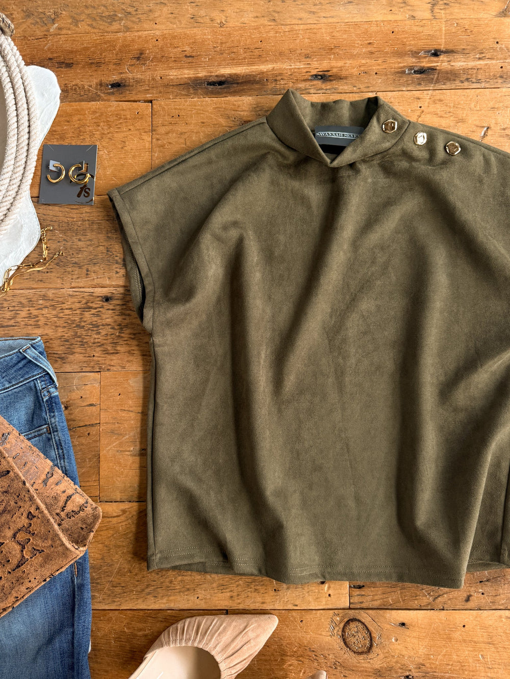 Seminole Olive Faux Suede Top by Savannah Sevens western life{&}style, featuring a relaxed fit and short sleeves with button details on the shoulder, is laid flat on a wooden surface. Styled nearby are blue jeans, a cork wallet, gold earrings, rope, and nude pointed-toe shoes.