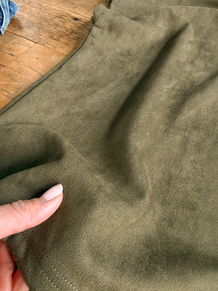 A hand with manicured nails pinches the corner of the Savannah Sevens western life{&}style Seminole Olive Faux Suede Top, with a wooden surface and part of a blue fabric visible in the background.