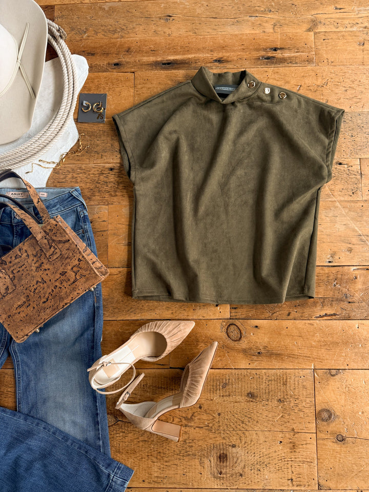 Flat lay of a casual outfit featuring the Seminole Olive Faux Suede Top by Savannah Sevens western life{&}style, paired with blue jeans, beige heels, a cork-textured handbag, beige hat, gold earrings, and a cream rope belt on a wooden floor.