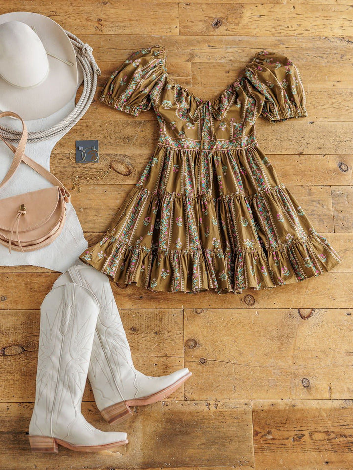 A flat lay of the Senoia Floral Puff Sleeve Dress by Savannah Sevens western life{&}style, paired with white cowboy boots, a beige hat, a tan crossbody purse, and gold earrings, displayed on a wooden floor.