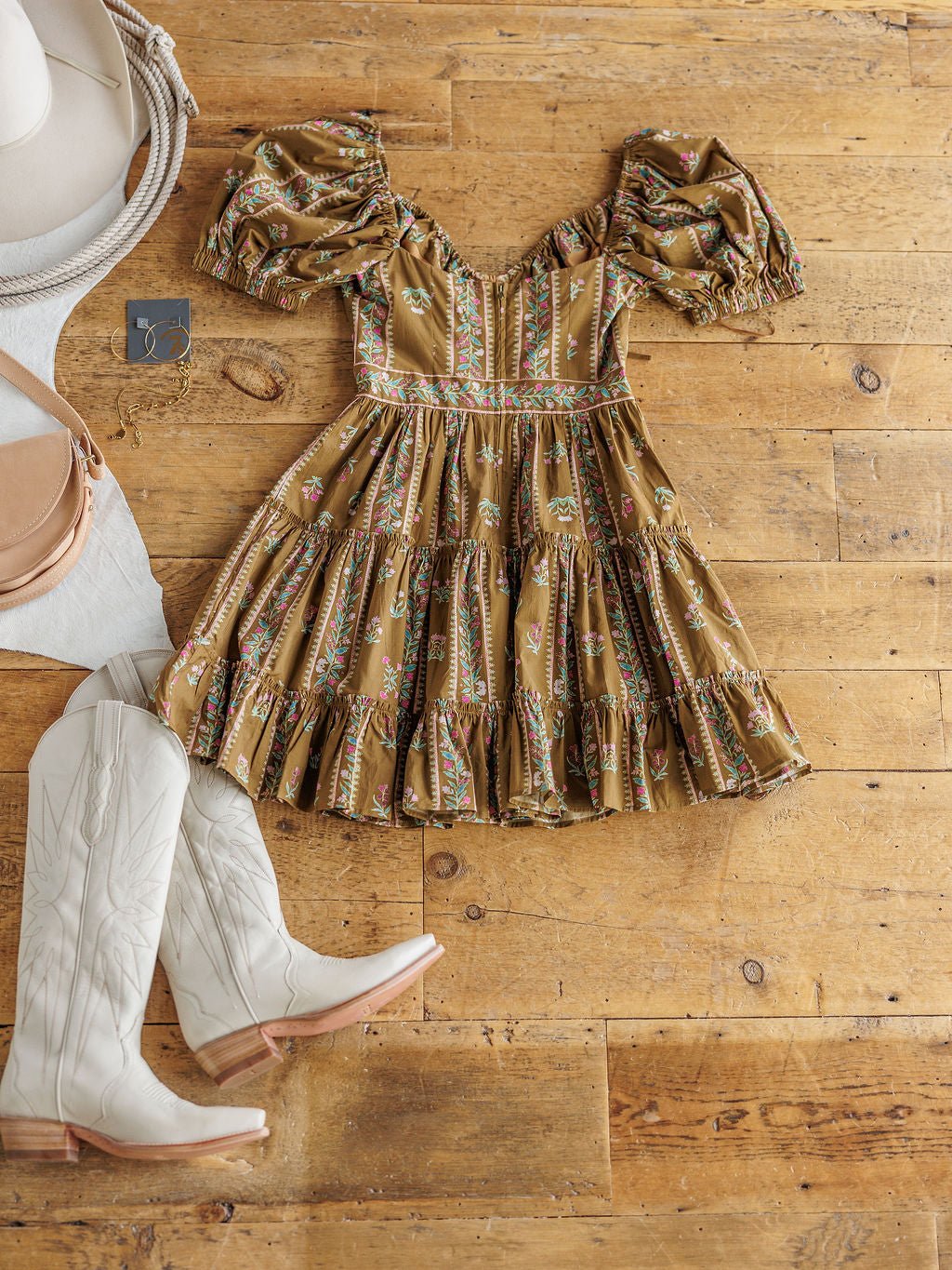 The Senoia Floral Puff Sleeve Dress by Savannah Sevens western life{&}style in brown is styled on a wooden floor with white cowboy boots, a round beige bag, a white hat, and gold earrings.