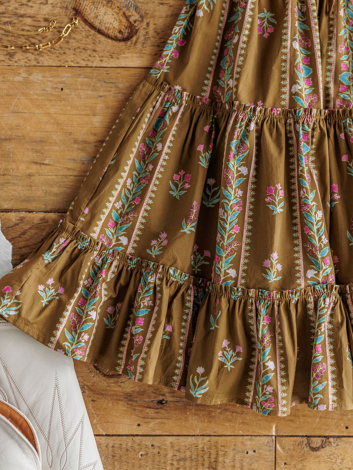 A close-up of the Senoia Floral Puff Sleeve Dress by Savannah Sevens western life{&}style, a cotton dress with green, pink, and blue floral embroidery, shown on wood next to a white embroidered boot and gold chain.