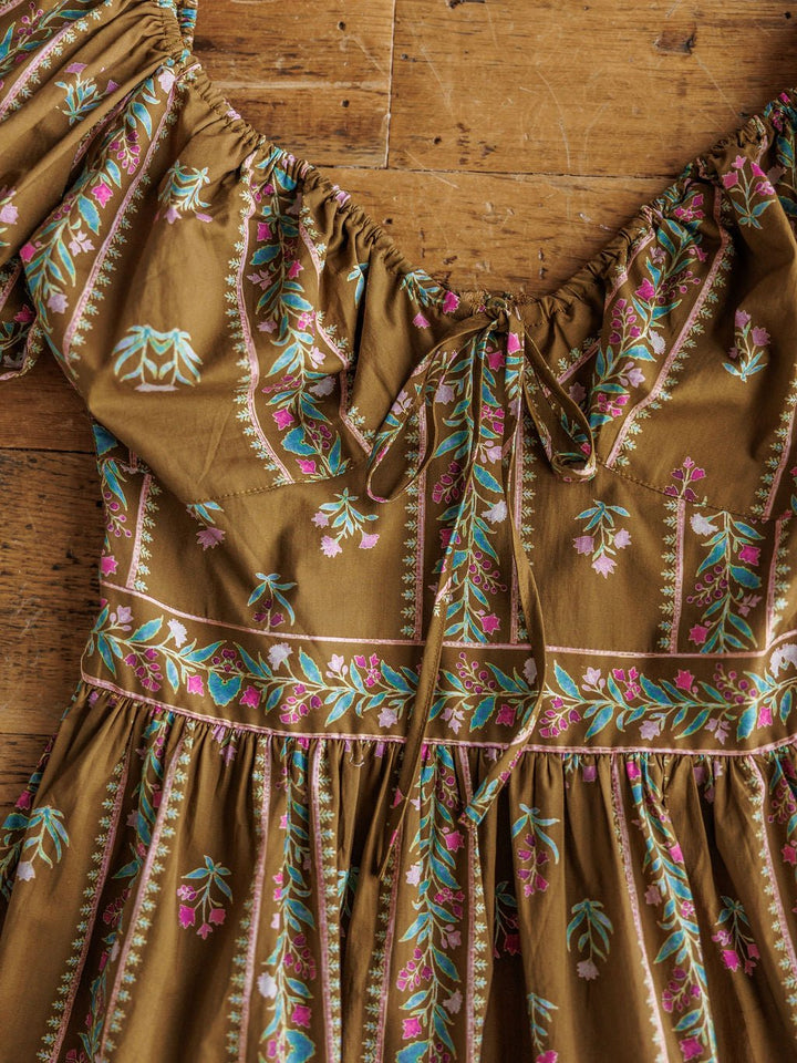 The Senoia Floral Puff Sleeve Dress by Savannah Sevens western life{&}style—a cotton dress in brown with green, pink, and white floral print, gathered tie neckline, and puffed sleeves—is displayed on a wooden surface.