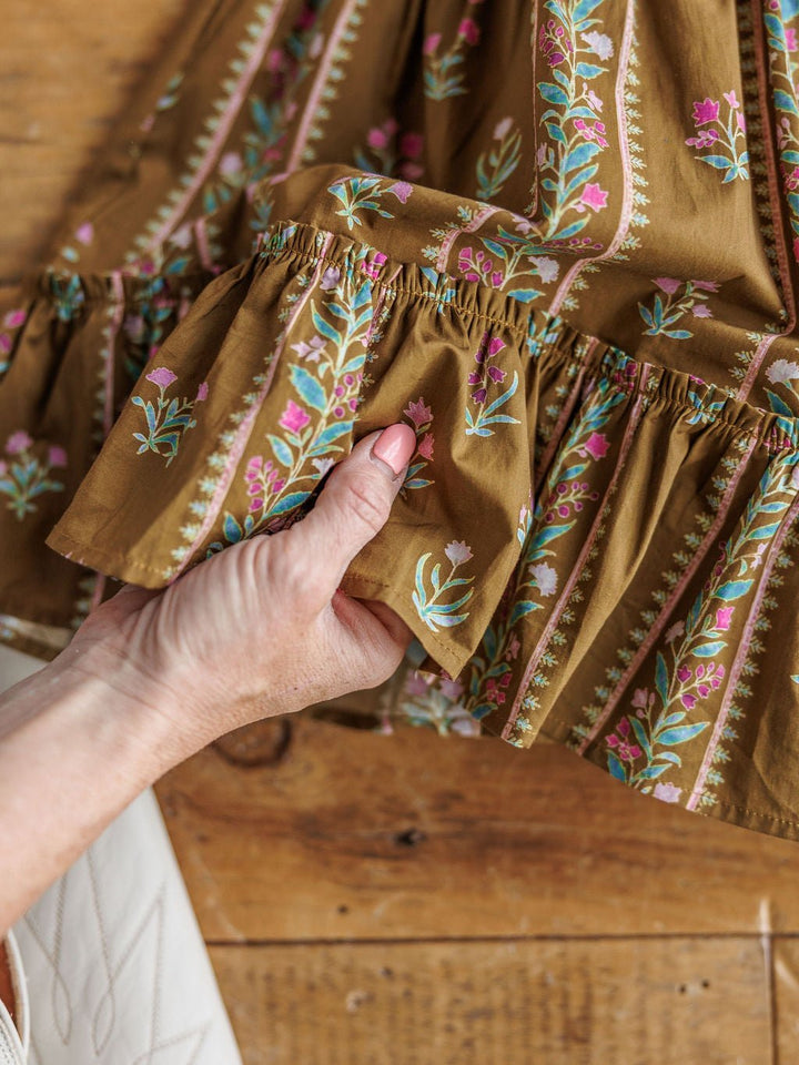 A hand with pink nail polish holds the ruffled hem of the Senoia Floral Puff Sleeve Dress by Savannah Sevens western life{&}style—a brown cotton dress with vertical floral stripes—set against a wooden background.