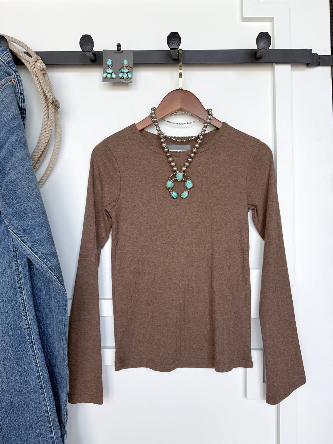 The Sentinel Mocha Knit Top by Savannah Sevens western life{&}style hangs on a wooden hanger. Turquoise jewelry—a necklace and earrings—are displayed with the top, while blue jeans are draped nearby on a black wall rack.