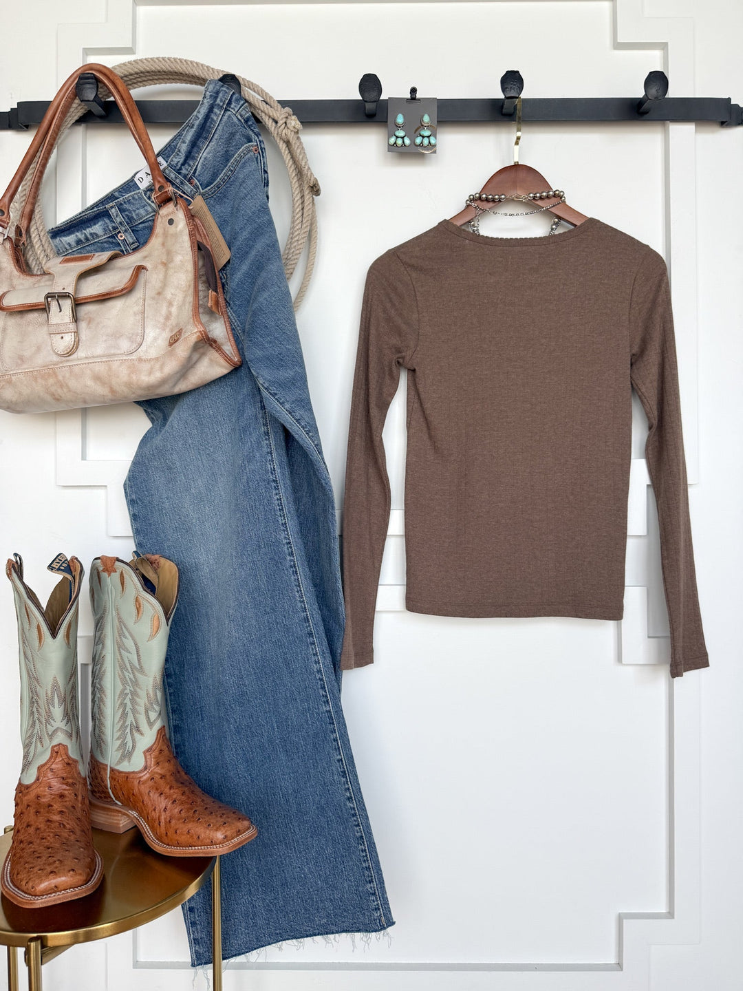 Savannah Sevens western life{&}style flat lay: Sentinel Mocha Knit Top, blue jeans, tan leather purse, turquoise cowboy boots with tan details, coiled rope, and turquoise earrings—all arranged against a white wall with black hooks.