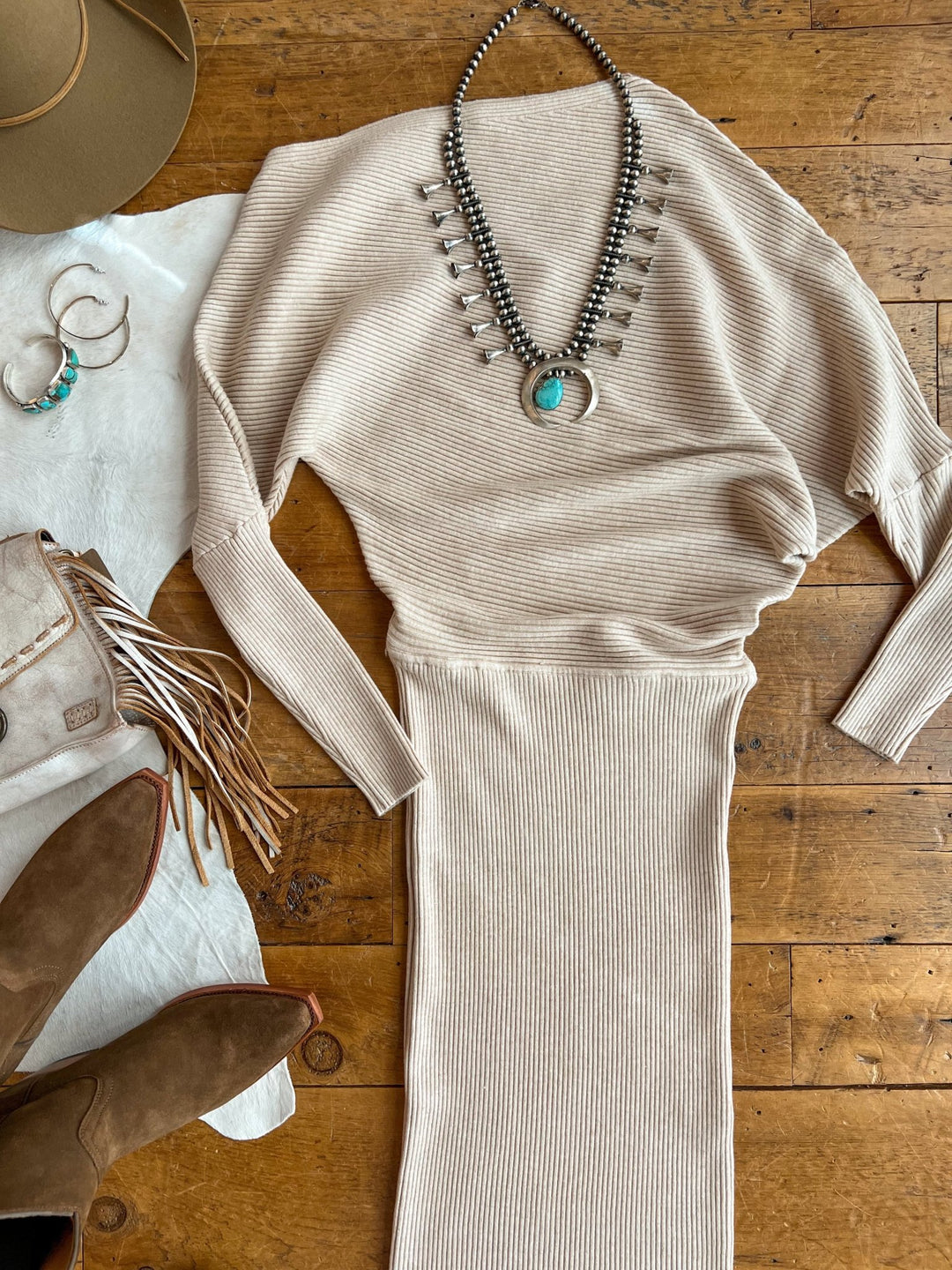The Shawnee Hills Oatmeal Ribbed Long Sleeve Dress by Savannah Sevens western life{&}style is styled with a chunky silver and turquoise necklace, brown boots, fringed bag, tan hat, and turquoise jewelry on a wooden floor.