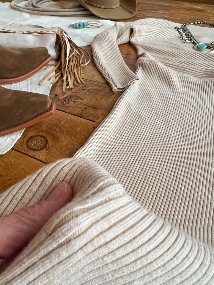 A close-up of the Shawnee Hills Oatmeal Ribbed Long Sleeve Dress by Savannah Sevens western life{&}style, laid out with brown boots, a tan hat, turquoise necklace, and fringe accessories on wood flooring. A hand holds the dress fabric in front.