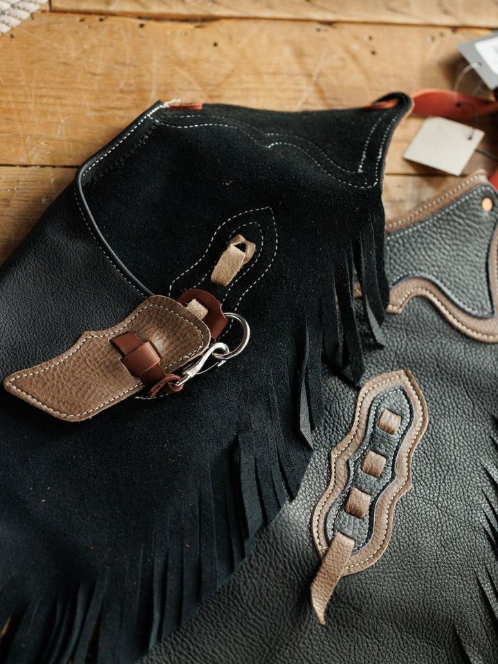 Close-up of Sheridan Chinks {Youth} by Savannah Sevens western life{&}style, featuring fringe, brown accents, detailed stitching, decorative cutouts, and an attached small leather sheath on a wooden surface. Adjustable for cowkids.