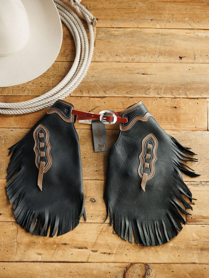 The Sheridan Chinks {Youth} by Savannah Sevens western life{&}style feature black leather with brown designs and fringe, shown with a coiled rope and white cowboy hat—ideal handmade chinks for youth needing an adjustable fit.