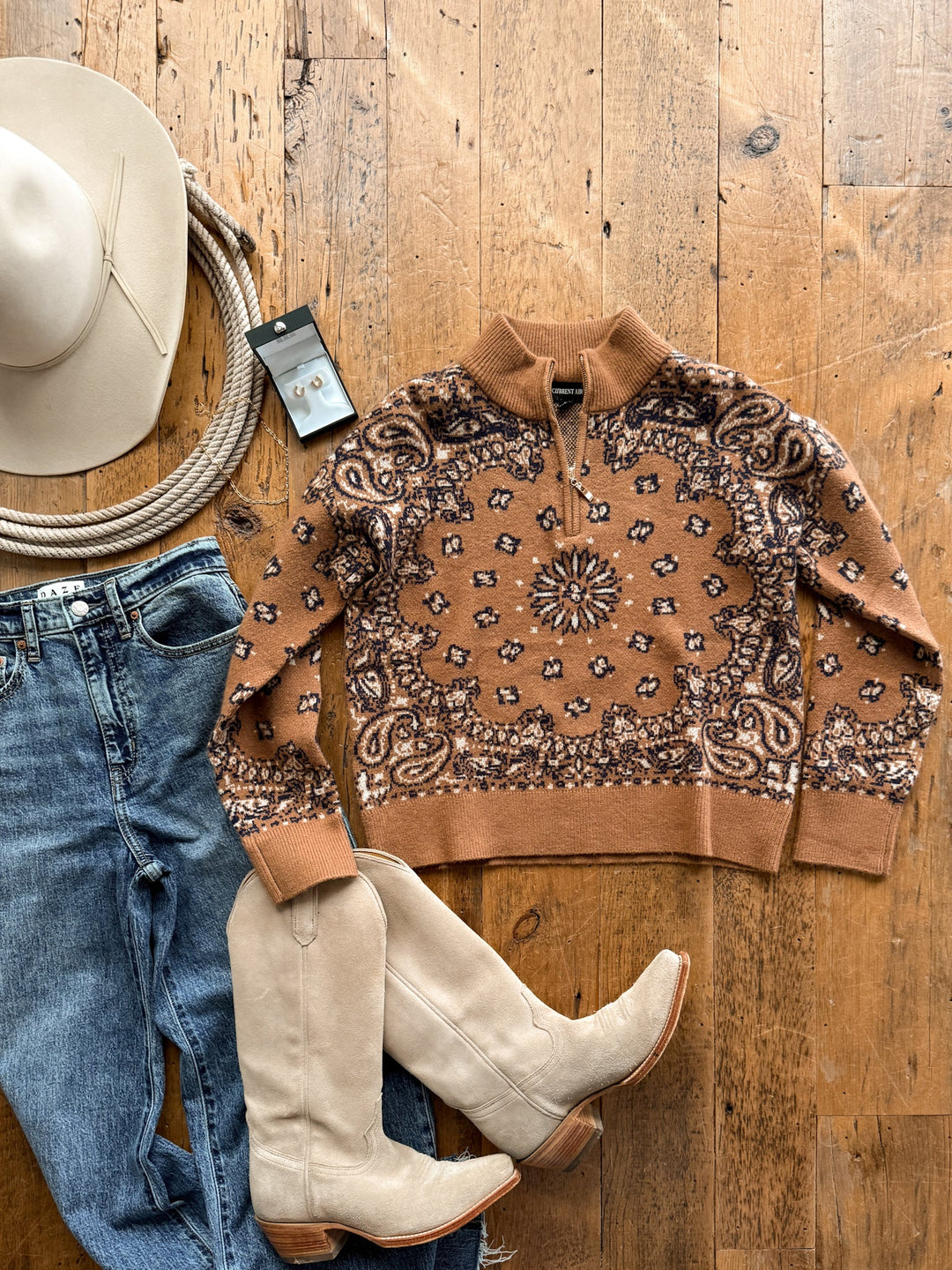 A Shiloh Caramel Paisley Quarter Zip Sweater by Savannah Sevens western life&style laid out with blue jeans, cream-colored western boots, a cowboy hat, a coiled rope, and a pair of earrings.
