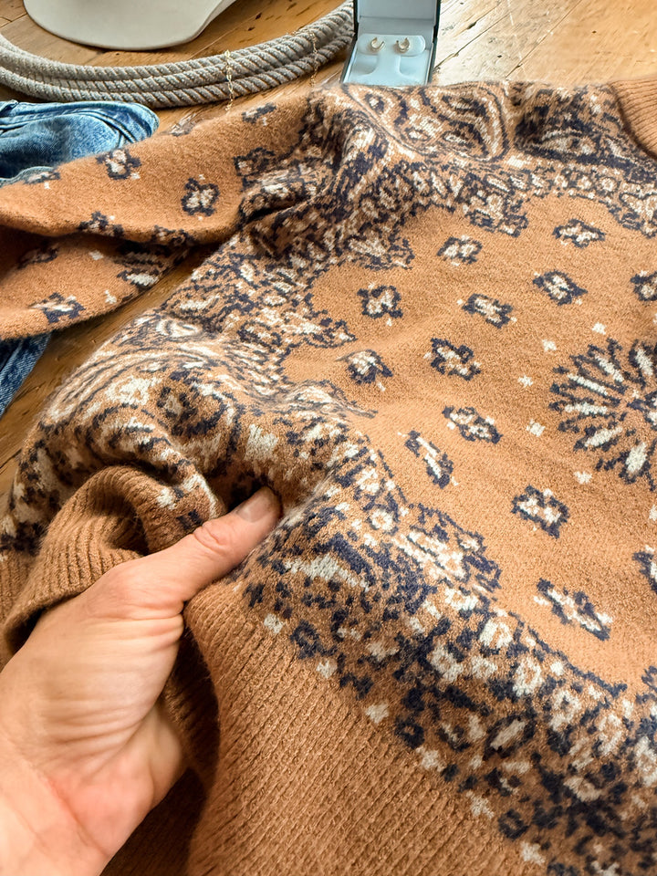 A hand holds a corner of a tan woven knit Shiloh Caramel Paisley Quarter Zip Sweater featuring a dark brown and cream paisley pattern.