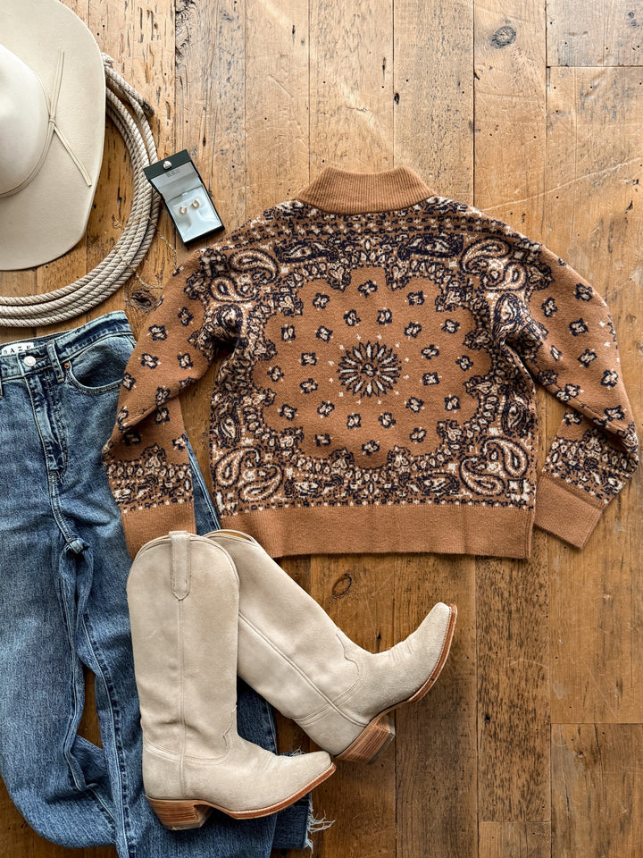 Shiloh Caramel Paisley Quarter Zip Sweater paired with blue jeans, cream cowboy boots, a cowboy hat, a rope, and small stud earrings laid out on a wooden surface.