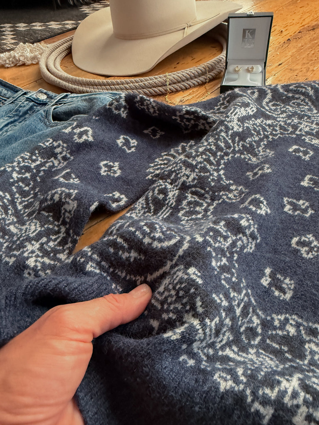 A hand holds a navy blue woven knit Shiloh Denim Paisley Quarter Zip Sweater by Savannah Sevens western life&style, lying on a wooden floor near jeans, a cowboy hat, and earrings.
