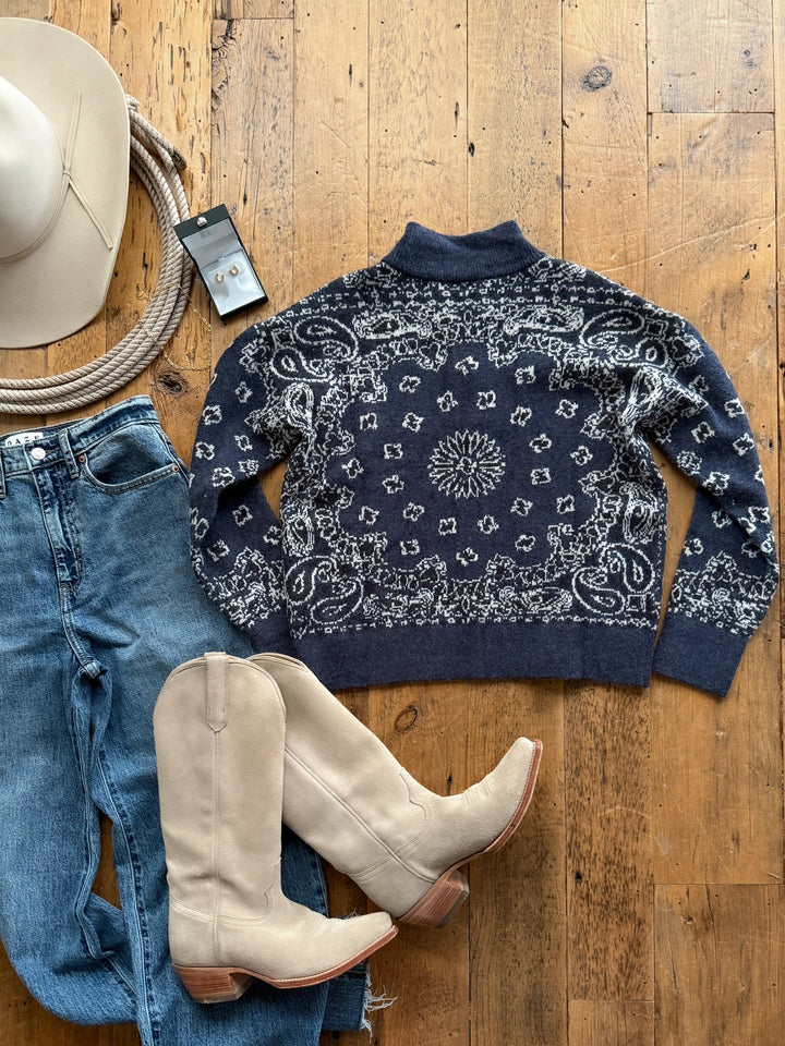 A Shiloh Denim Paisley Quarter Zip Sweater in navy blue with a white paisley pattern, denim jeans, light-colored western boots, a felt hat, a coiled rope, and a pair of earrings laid out on a wooden floor.