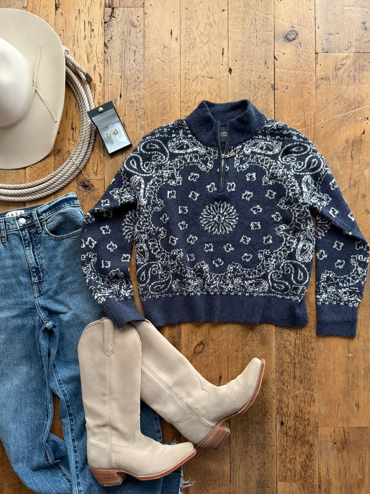 A navy blue paisley print Shiloh Denim Paisley Quarter Zip Sweater by Savannah Sevens western life&style laid on a wooden floor with blue jeans, tan western boots, a cowboy hat, a coiled rope, and a pair of stud earrings.