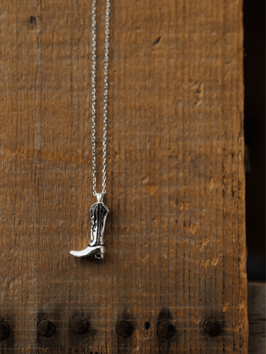 The Silver Cowgirl Boot Necklace by Savannah Sevens western life{&}style hangs against a rustic wooden background, showcasing its western charm with visible grain and metal studs.