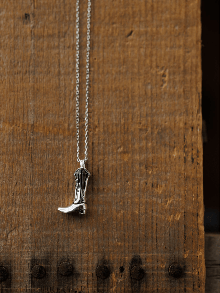 The Silver Cowgirl Boot Necklace by Savannah Sevens western life{&}style hangs against a rustic wooden background, showcasing its western charm with visible grain and metal studs.