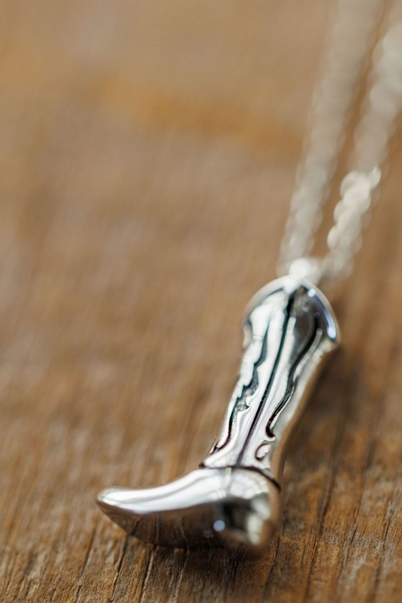 A close-up of the Silver Cowgirl Boot Necklace by Savannah Sevens western life{&}style, showcasing its decorative engravings and fine detailing on a chain, with the boot in sharp focus against a softly blurred wooden background.