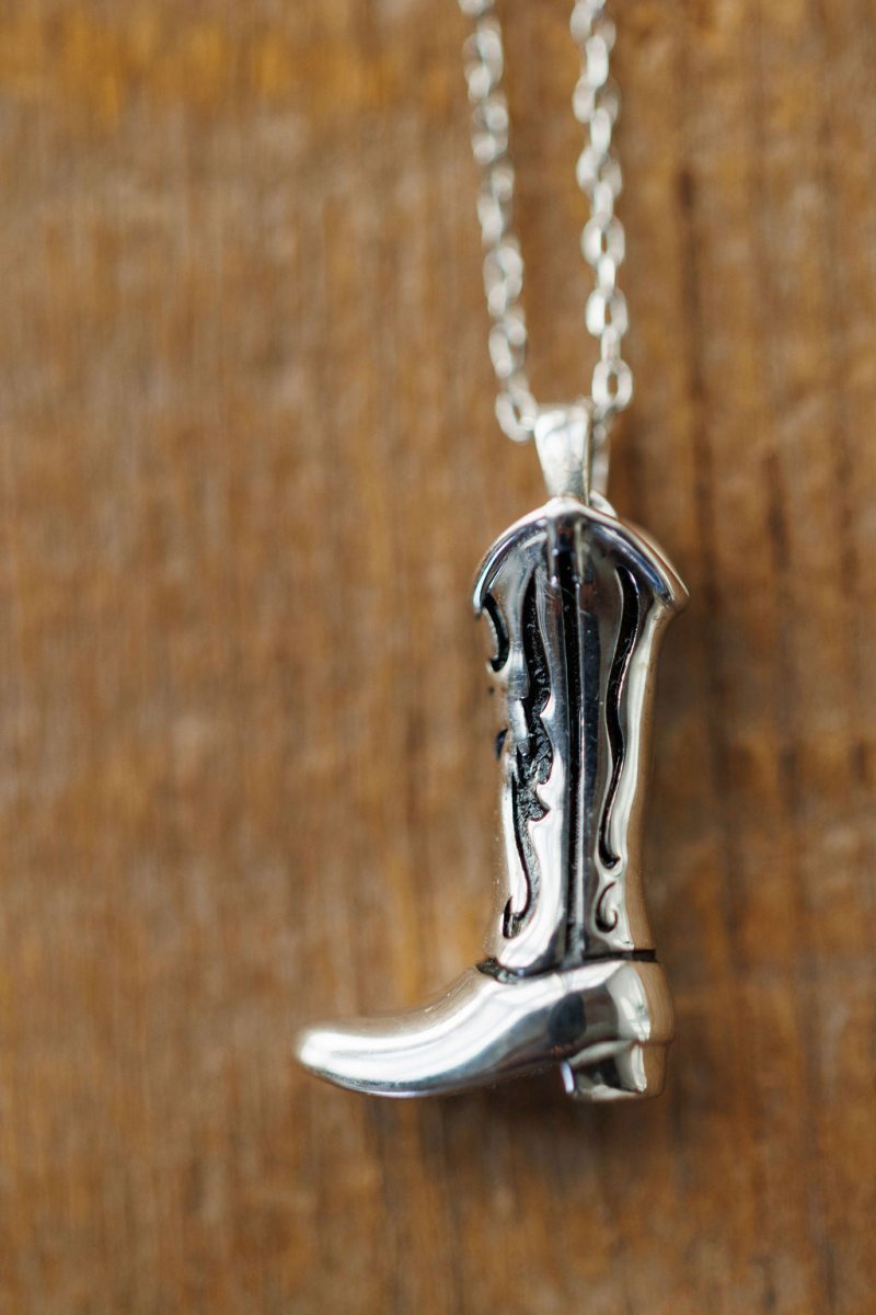 The Silver Cowgirl Boot Necklace by Savannah Sevens western life{&}style features engraved swirl designs, shown against a wooden background for a touch of classic western charm.