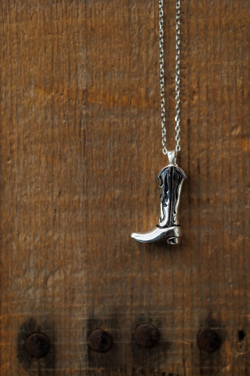The Silver Cowgirl Boot Necklace by Savannah Sevens western life{&}style is displayed against a rustic wooden background with visible grain.