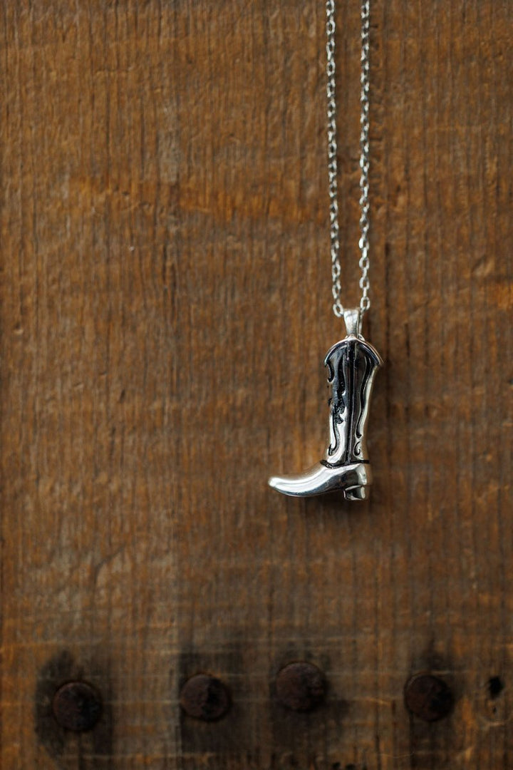 The Silver Cowgirl Boot Necklace by Savannah Sevens western life{&}style is displayed against a rustic wooden background with visible grain.