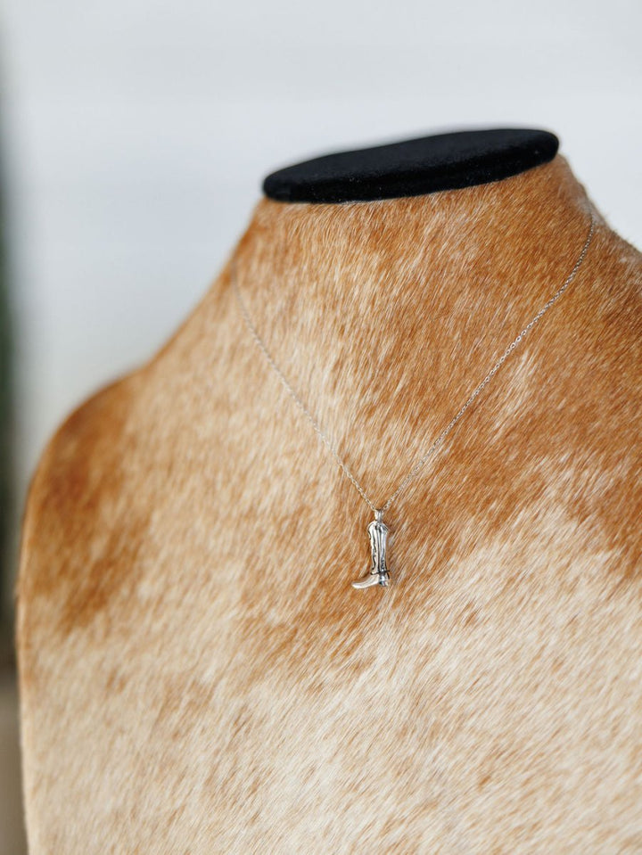 The Silver Cowgirl Boot Necklace by Savannah Sevens western life{&}style features a dainty boot pendant, shown on a brown and white cowhide jewelry bust.