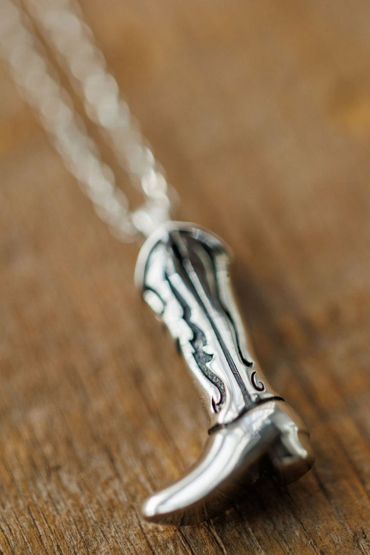 Close-up of the Silver Cowgirl Boot Necklace by Savannah Sevens western life{&}style, featuring engraved decorative patterns and hanging from a chain on a wooden surface.