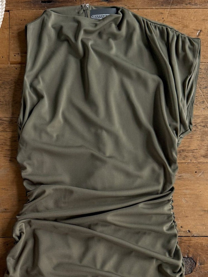 The Sinton Olive Bodycon Dress by Savannah Sevens western life{&}style, featuring a high neckline and short sleeves, is displayed flat on a wooden surface. This ruched dress showcases draped olive green fabric with gathered detailing along the sides.