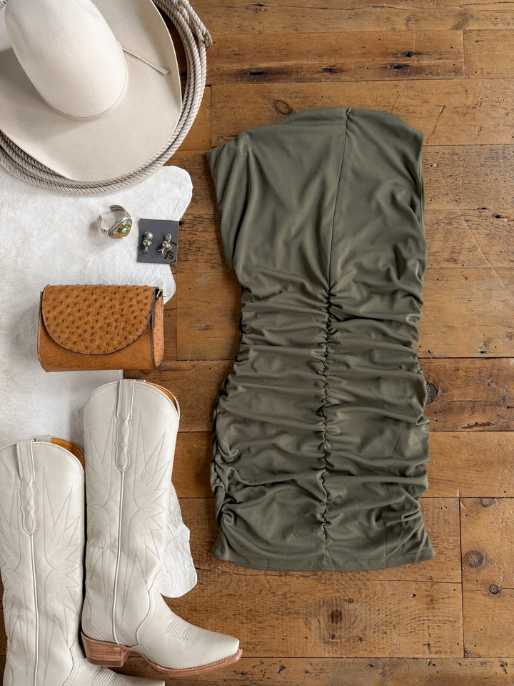 Flat lay of the Sinton Olive Bodycon Dress by Savannah Sevens western life{&}style, styled with white cowboy boots, a tan clutch, silver jewelry, and a white cowboy hat on a wooden floor beside a white fur rug. This chic ensemble is perfect for making a stylish statement.