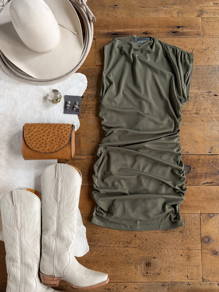 A flat lay featuring the Sinton Olive Bodycon Dress by Savannah Sevens western life{&}style, paired with ivory cowboy boots, a cream hat, a tan wallet, and silver jewelry—all arranged on a wooden floor with a white fur rug.