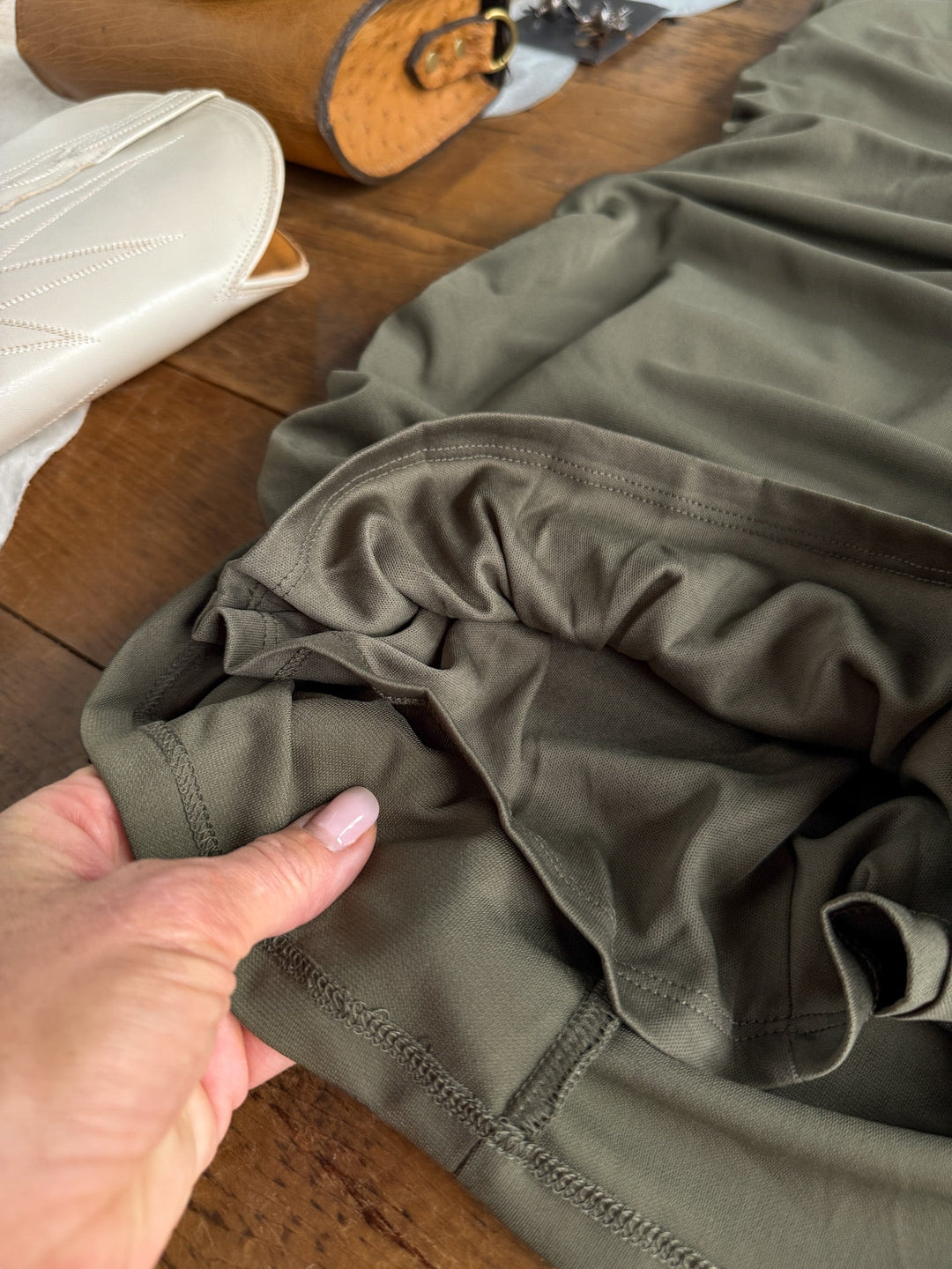 A hand holds the waistband of the Sinton Olive Bodycon Dress from Savannah Sevens western life{&}style, laid on a wooden surface with a white purse and a brown bag partially visible in the background, evoking its signature sleek style.