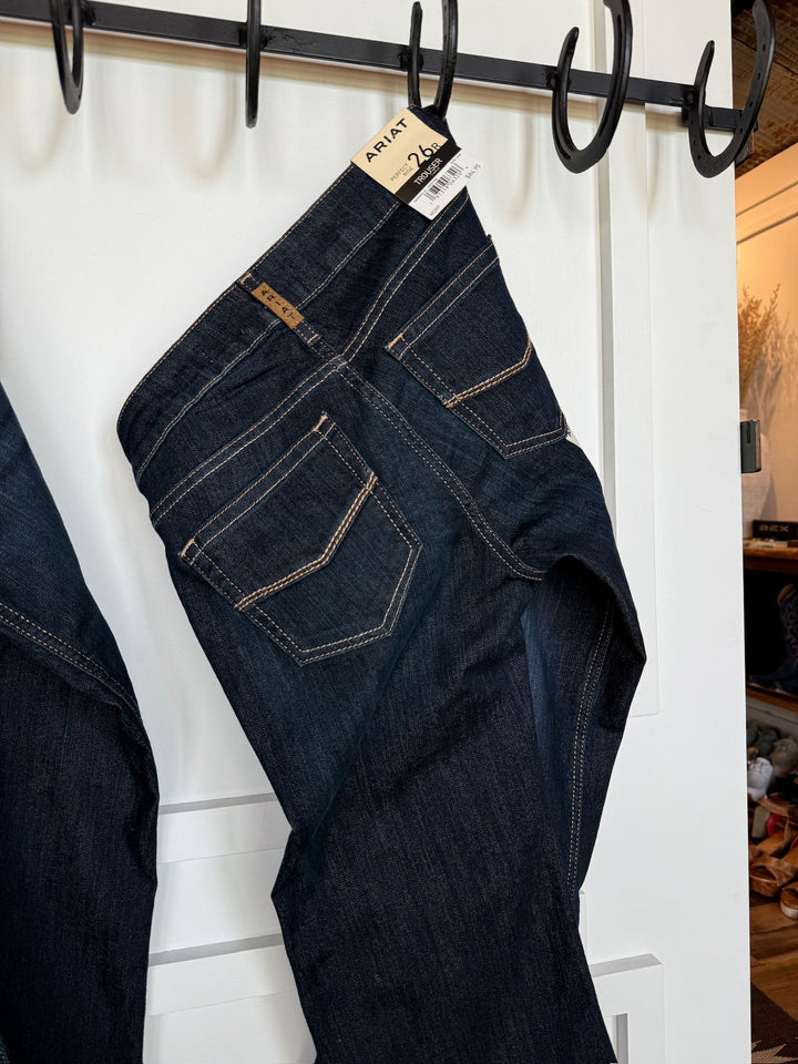 A pair of The Slade Perfect Rise Trouser Jeans by Savannah Sevens western life{&}style hangs from a black hook rack on a white door, featuring tan stitching, visible back pockets, and a price tag on the waistband.