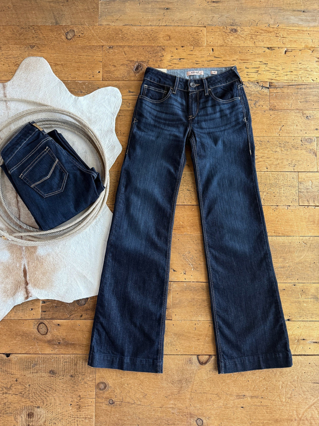 A pair of The Slade Perfect Rise Trouser Jeans by Savannah Sevens western life{&}style lies flat on a wooden floor beside folded jeans and a white cowhide rug, highlighting the classic bootcut style.
