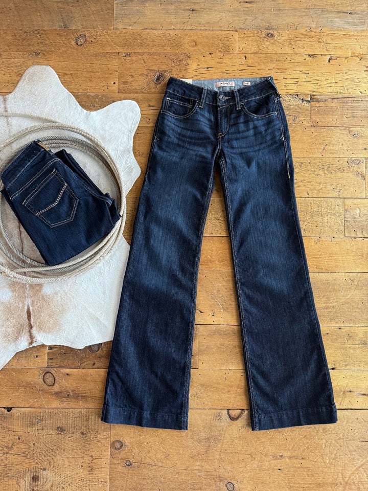 A pair of The Slade Perfect Rise Trouser Jeans by Savannah Sevens western life{&}style lies flat on a wooden floor beside folded jeans and a white cowhide rug, highlighting the classic bootcut style.