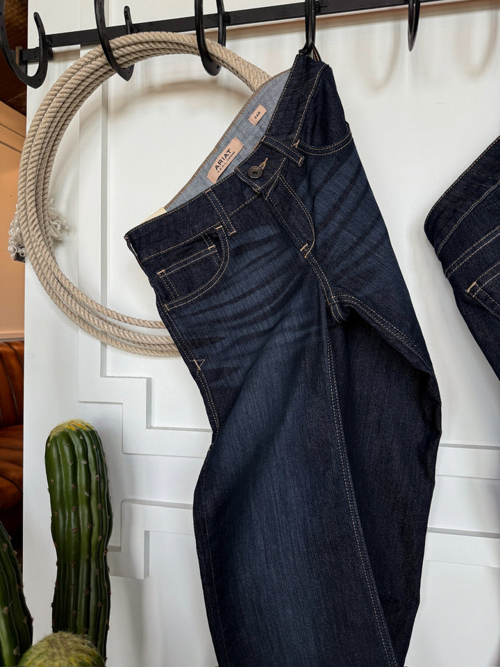 The Slade Perfect Rise Trouser Jean by Savannah Sevens western life{&}style hangs by the waist from a thick rope against a white wall, with a green cactus partly visible in the lower left corner.