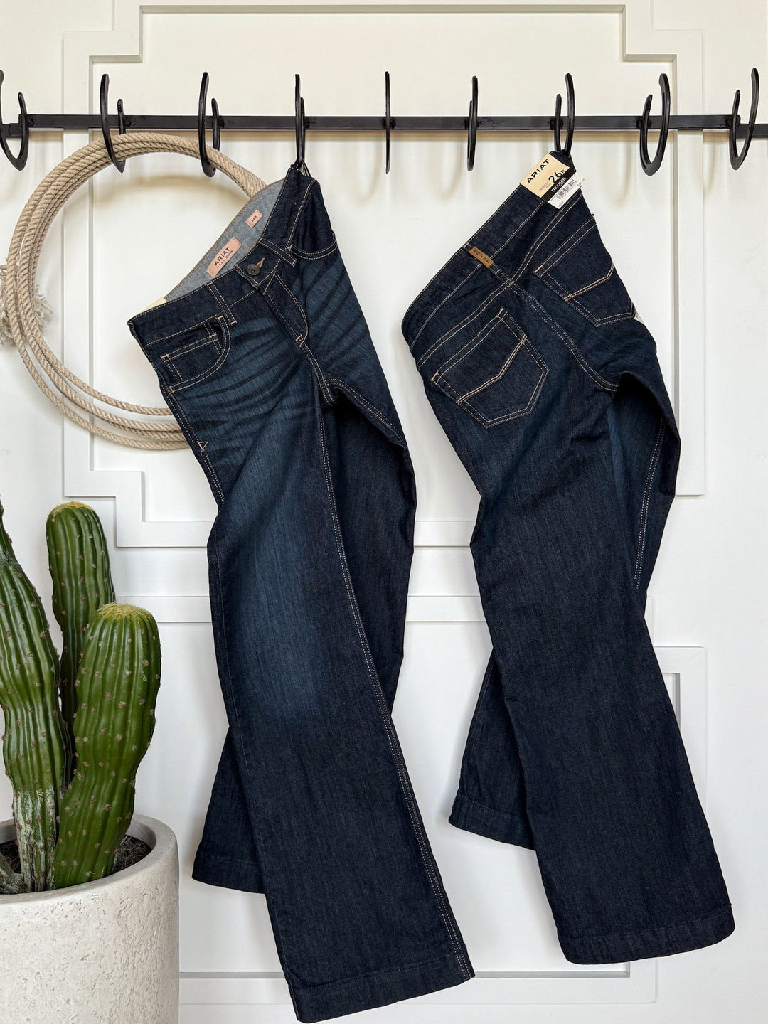 Two pairs of Savannah Sevens western life{&}style The Slade Perfect Rise Trouser Jeans hang side by side on black hooks on a white door, with a potted cactus nearby and a circular rope hanging to the left.