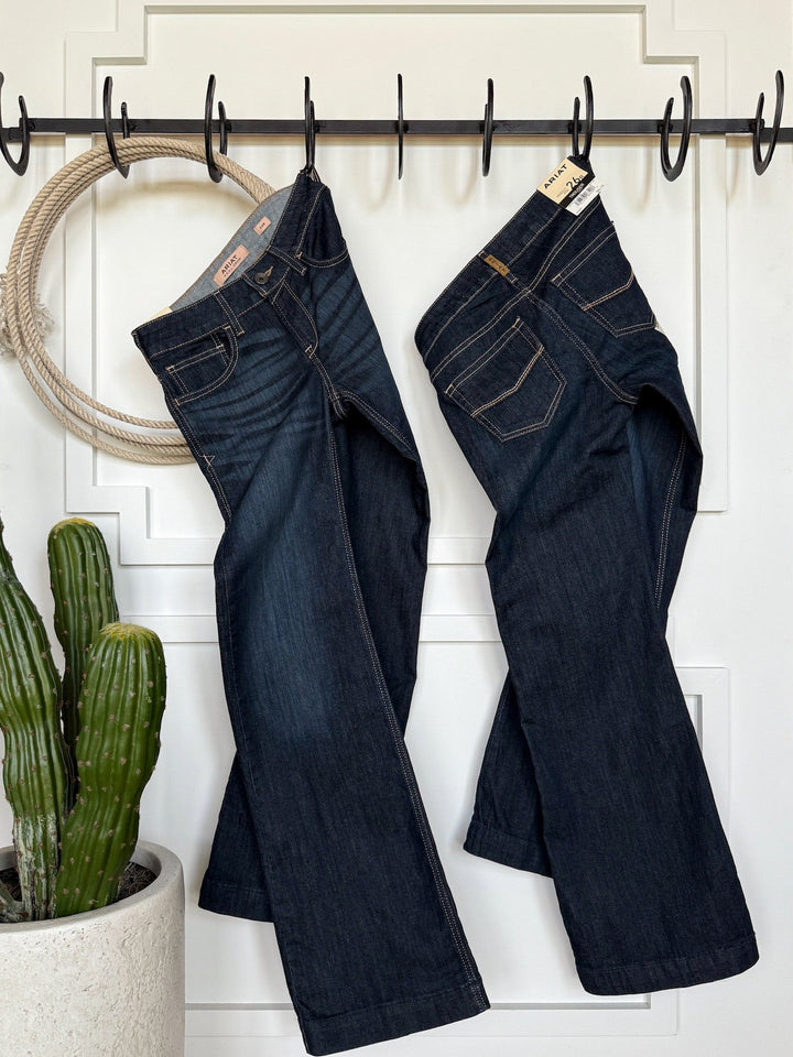 Two pairs of Savannah Sevens western life{&}style The Slade Perfect Rise Trouser Jeans hang side by side on black hooks on a white door, with a potted cactus nearby and a circular rope hanging to the left.